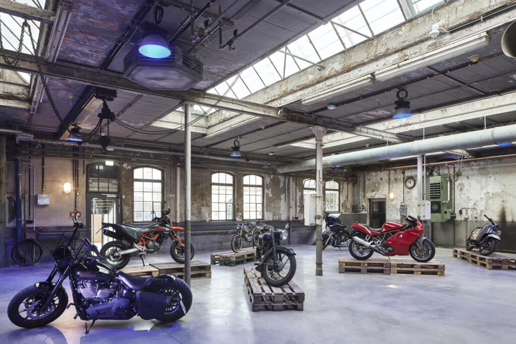 Großer, alter Industriehallenraum mit sechs verschiedenen Motorrädern und einem Fahrrad, die auf Holzpaletten ausgestellt sind, sichtbare Rohre und verwitterte Wände mit großen Fenstern und Deckenfenstern, die von blauem Licht beleuchtet werden