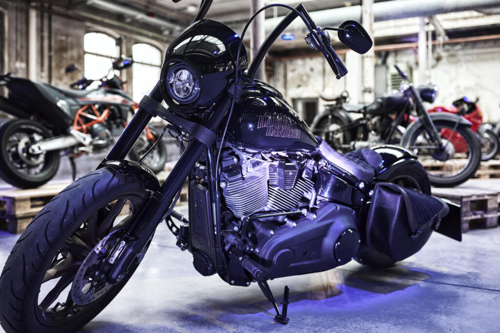 Schwarzes Harley-Davidson-Motorrad mit markantem Frontscheinwerfer und Leder-Satteltasche in einer Industriehalle mit weiteren Motorrädern im Hintergrund