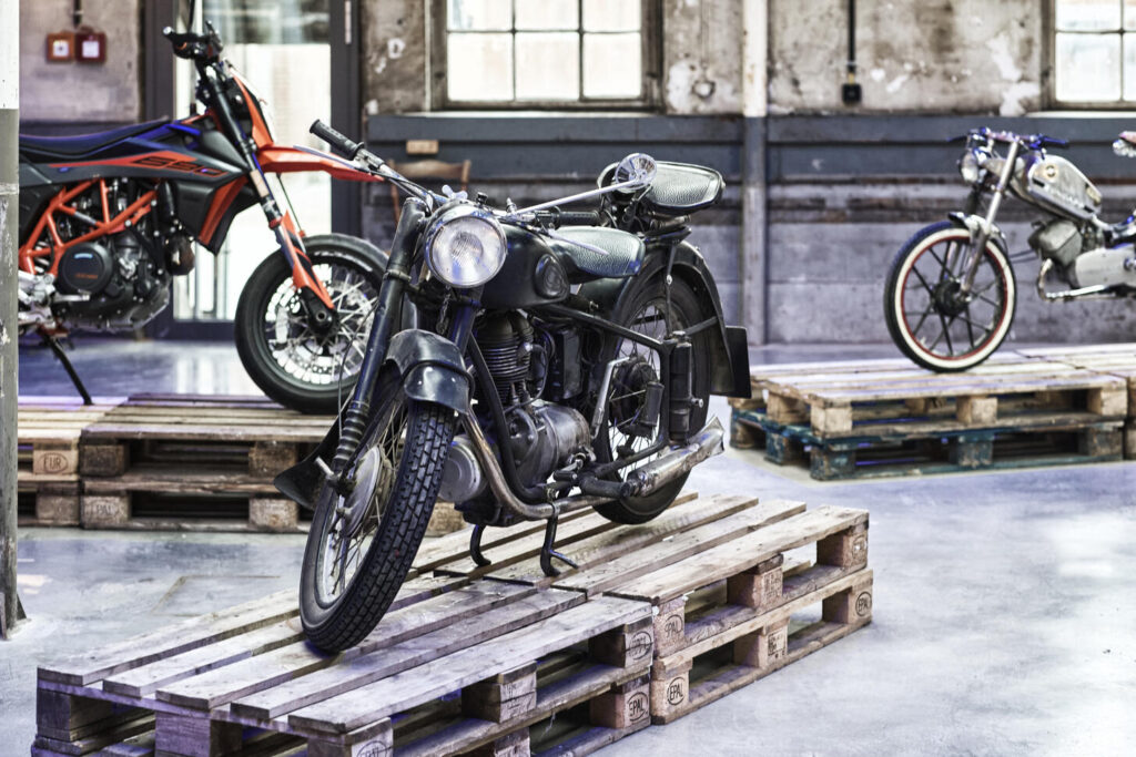 Drei Motorräder unterschiedlicher Stile stehend auf Holzpaletten in einem hellen, industriellen Innenraum mit groben Wänden und großen Fenstern.