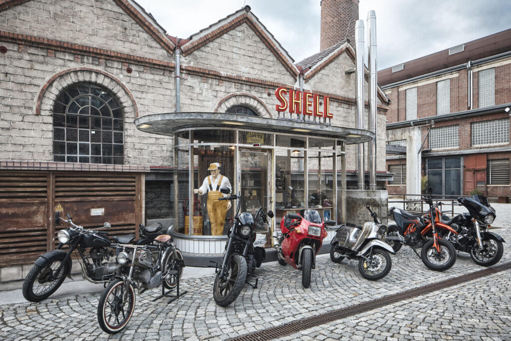 Mehrere Motorräder und Motorroller stehen vor einem historischen Gebäude mit dem Schild "SHELL" und einer Schaufensterpuppe in Arbeitskleidung in einer Glasvitrine