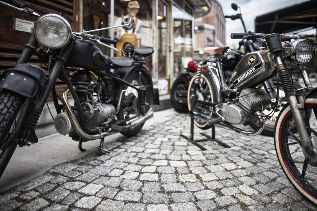 Zwei alte Motorräder auf gepflastertem Gehweg vor einem Geschäft, eines davon mit Zündapp-Logo, unscharfer Hintergrund mit Figur und Gebäuden
