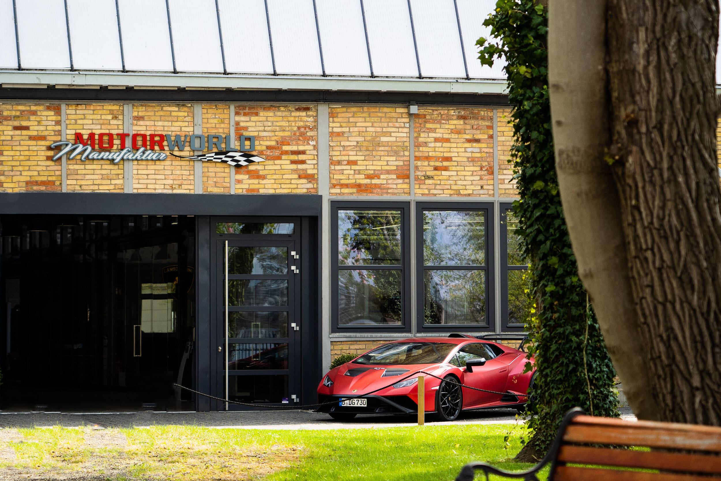 Roter Sportwagen vor einem Gebäude mit hellen Ziegeln und dem Schriftzug 'Motorworld Manufaktur', im Vordergrund Baumstämme, Efeu und eine Holzbank auf einer Grünfläche.