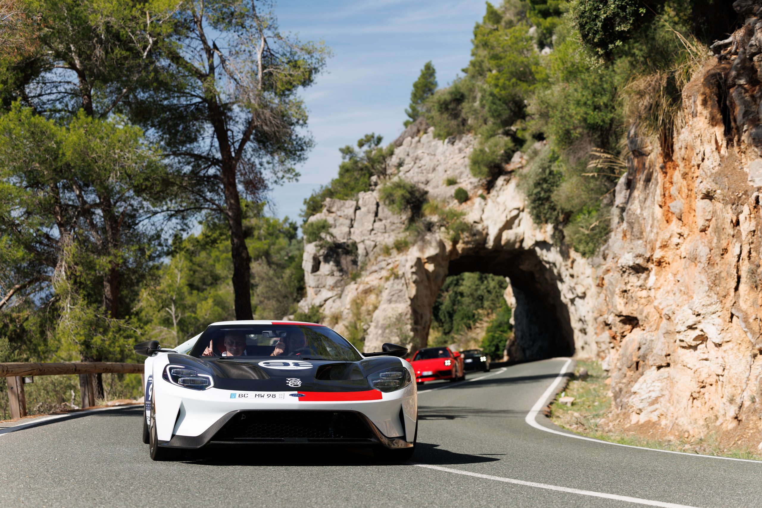 Vorderansicht eines weißen Sportwagens mit schwarzen und roten Akzenten, der auf einer kurvigen Bergstraße mit Felsformationen und Bäumen fährt, im Hintergrund weitere Sportwagen und ein natürlicher Felsdurchgang auf der Straße.