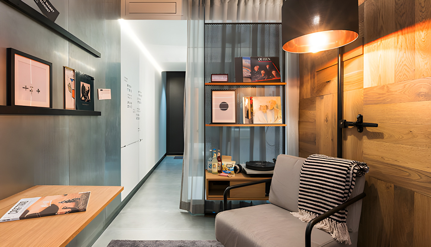 Moderner Wohnraum mit grauem Sofa, einer schwarz-weißen gestreiften Decke, hölzerner Wandverkleidung und einem stehendem Lampenschirm, links davon Regale mit Kunstwerken und Dekorationen sowie einem Flur mit heller Beleuchtung im Hintergrund.