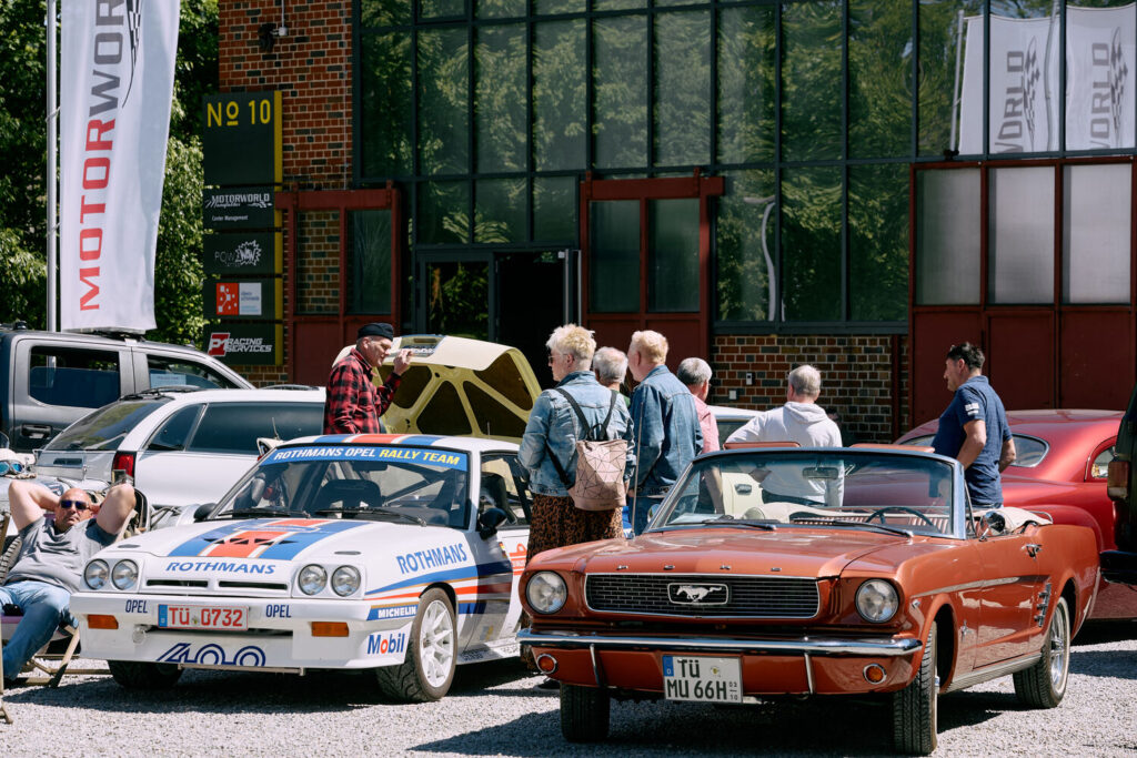 Mehrere Personen stehen um zwei klassische Autos, einen weißen Rothmans Opel Rallye Wagen mit geöffnetem Heck und einen roten Ford Mustang Cabrio, vor einem Gebäude mit großen Glasfenstern und Motorworld-Flaggen im Hintergrund.
