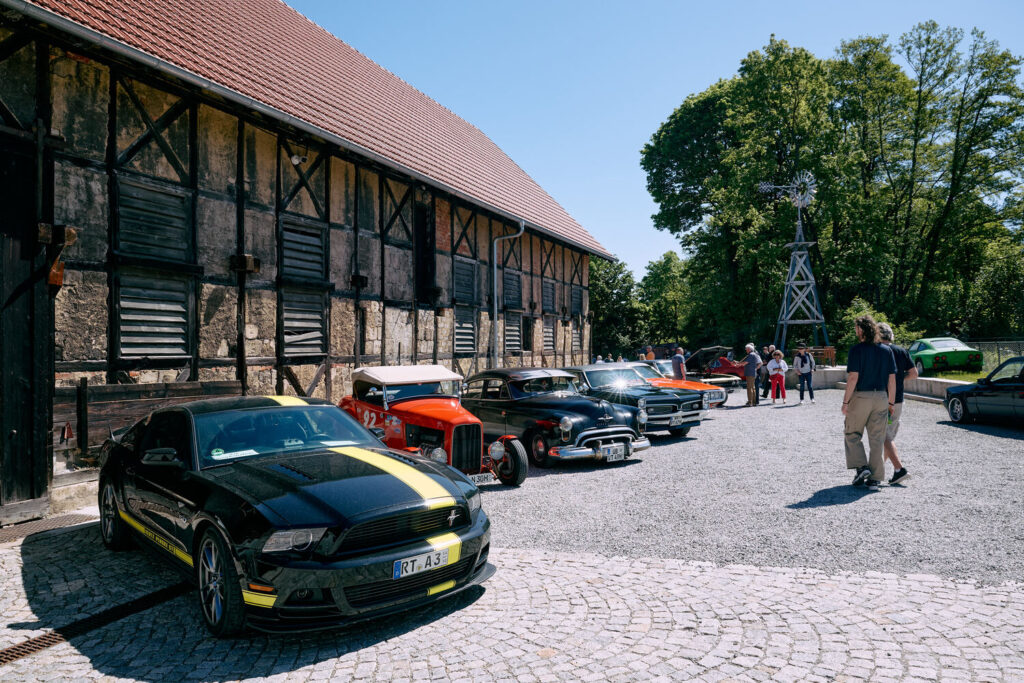 Mehrere Oldtimer-Autos in einer Reihe vor einem historischen Fachwerkgebäude mit rotem Dach und daneben Menschen, im Hintergrund Bäume und eine kleine Windmühle, alles unter klarem blauem Himmel an einem sonnigen Tag.