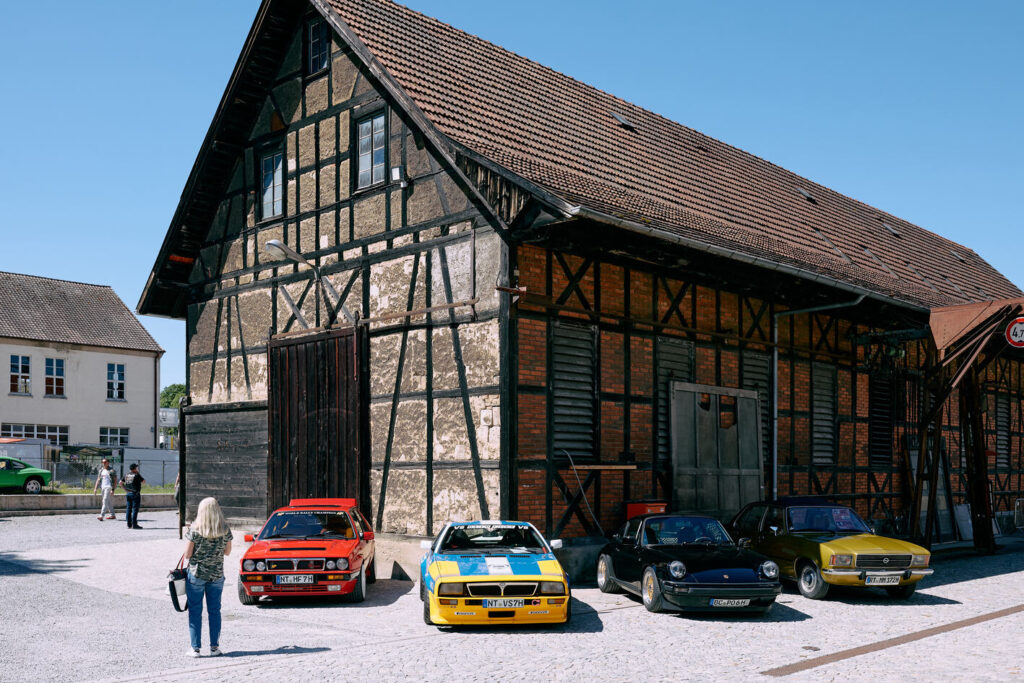 Vier Oldtimer-Autos in verschiedenen Farben vor einem traditionellen Fachwerkgebäude mit rotem Ziegeldach an einem sonnigen Tag