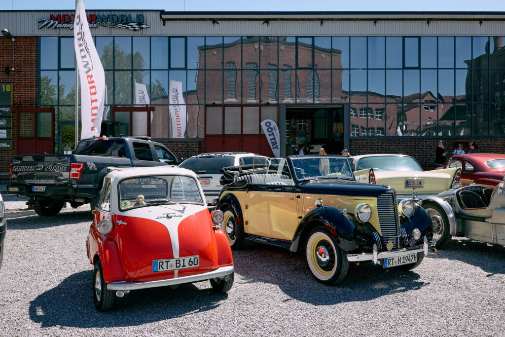 Parkplatz mit verschiedenen klassischen und modernen Autos vor einem Gebäude mit großen, spiegelnden Fenstern, die ein Backsteingebäude reflektieren, darunter ein rotes und weißes kleines Auto und ein gelb-schwarzes Cabrio.