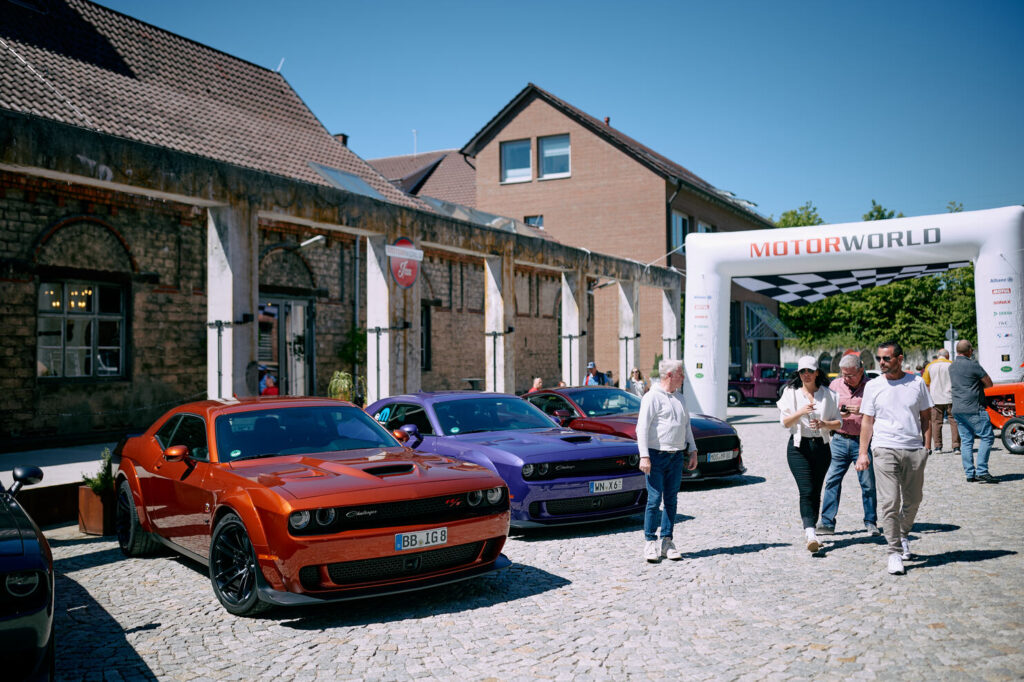 Drei auffällige Dodge Challenger Sportwagen in Orange, Lila und Dunkelrot stehen auf einem gepflasterten Platz vor einem alten Gebäude, daneben gehen mehrere Menschen an einem weißen Startbogen mit der Aufschrift ‚MOTORWORLD‘ vorbei unter klarem blauem Himmel.