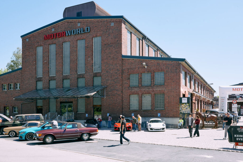Gebäude mit der Aufschrift «MOTORWORLD», davor mehrere klassische und moderne Autos sowie mehrere Menschen auf einem gepflasterten Platz bei sonnigem Wetter.
