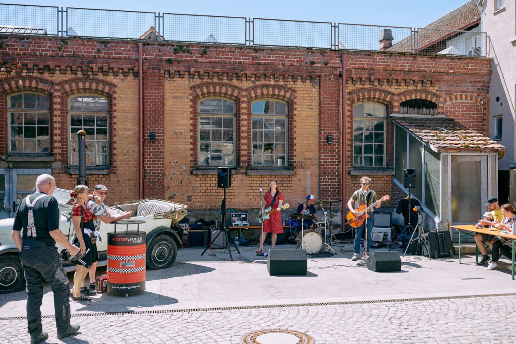 Drei Musiker spielen vor einer alten Ziegelfassade, eine Frau am Bass, ein Mann an der Gitarre und ein Schlagzeuger, während mehrere Menschen in der Sonne stehen und an einem Tisch sitzen, neben einem alten weißen Cabrio und einer roten Fass-Bühne mit der Aufschrift MOTORWORLD.