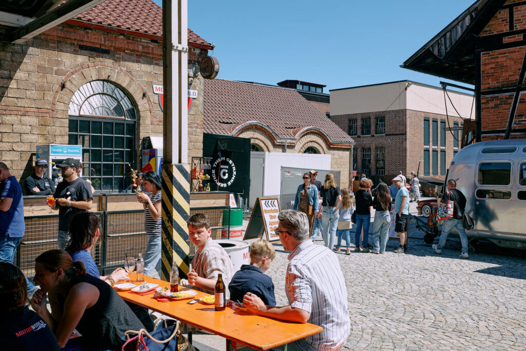 Menschen sitzen an einem langen Holztisch und essen bei sonnigem Wetter vor einem Gebäude mit Backsteinfassade, während weitere Personen in einer Schlange vor einem Getränkestand warten.