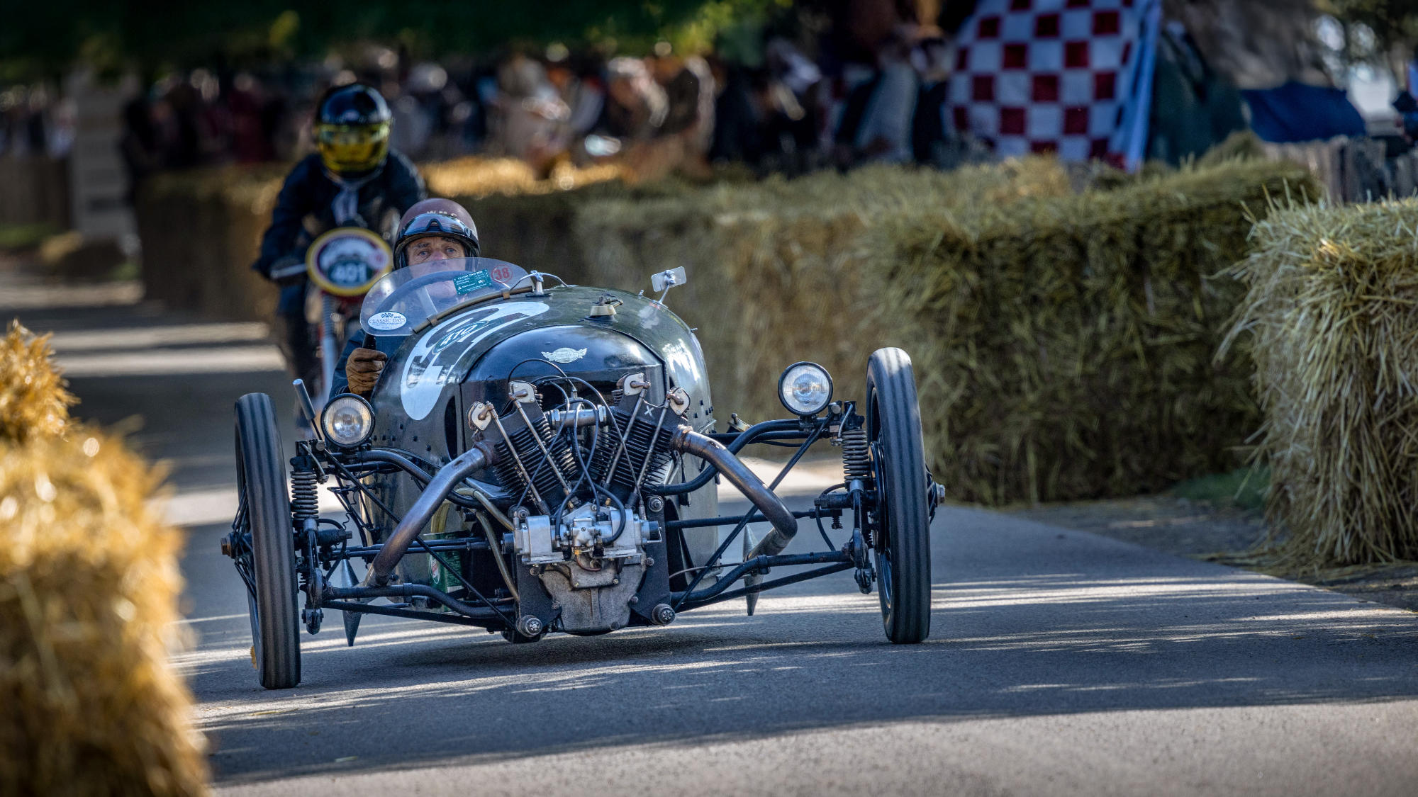 Historischer Rennwagen mit offenem Motor und Fahrer mit Helm fährt auf einer von Strohballen gesäumten Straße, im Hintergrund ein Motorradfahrer mit Helm.