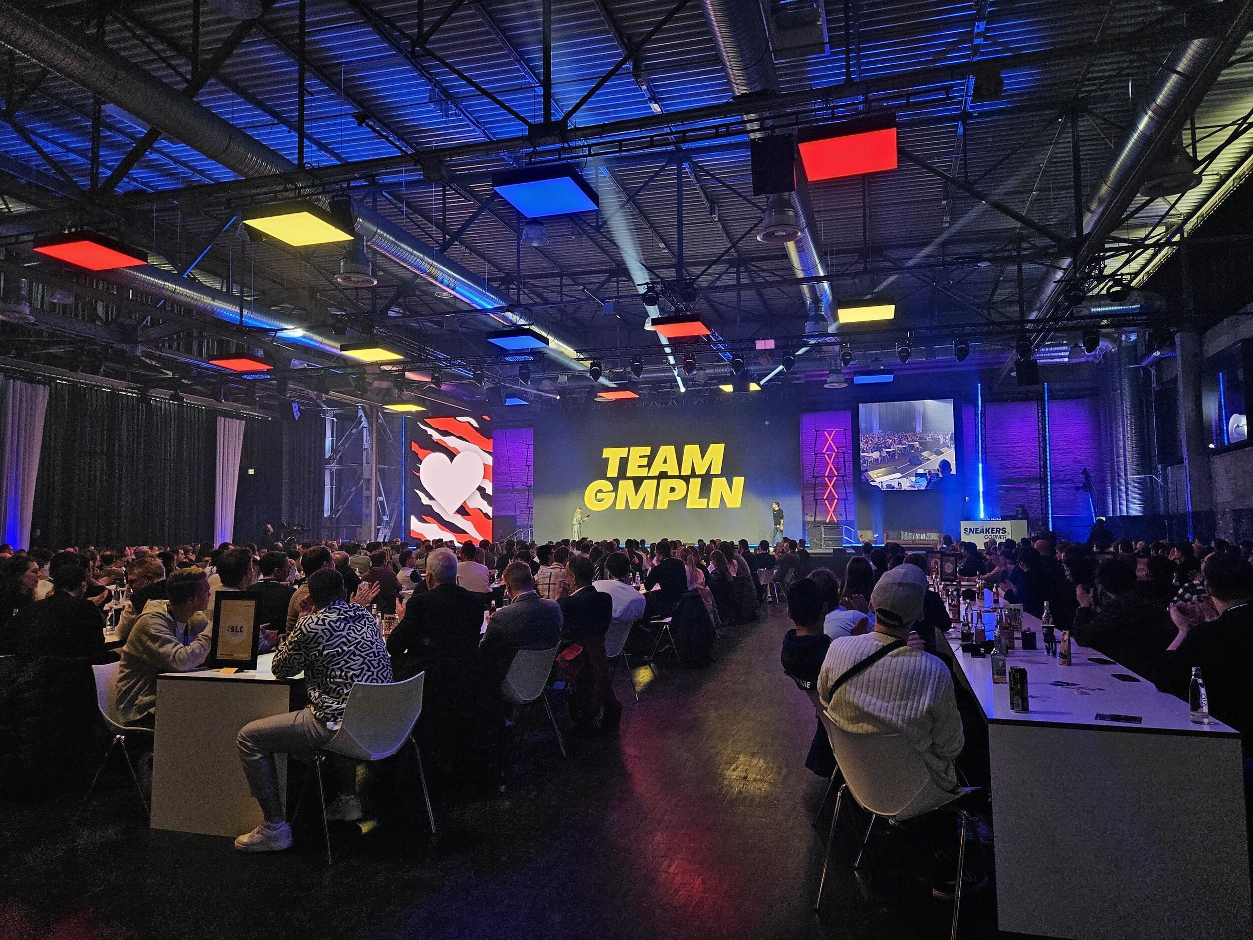 Vollbesetzter Konferenzraum mit Publikum, das zu einer beleuchteten Bühne mit der Aufschrift TEAM GMPLN schaut, umgeben von bunten Lichtern und Bildschirmen in einer Industriehalle.