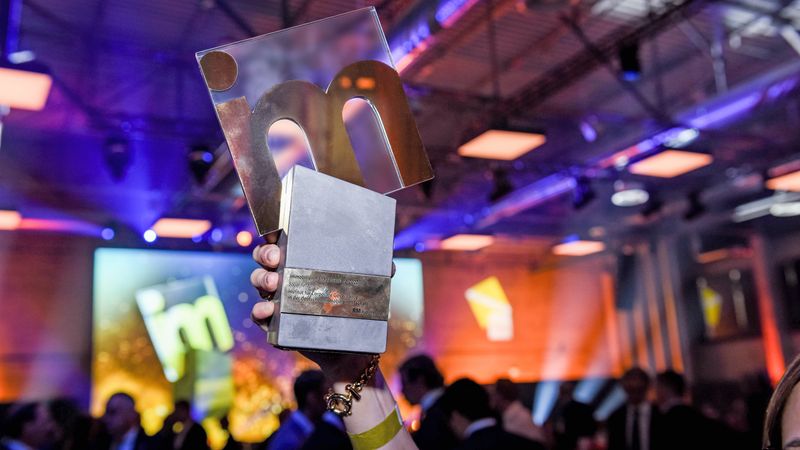 Nahaufnahme einer Hand, die eine moderne Gewinner-Trophäe mit einem großen, durchbrochenen Buchstaben "m" hält, vor einem unscharfen Hintergrund eines Eventsaals mit Menschen und bunten Lichtern.