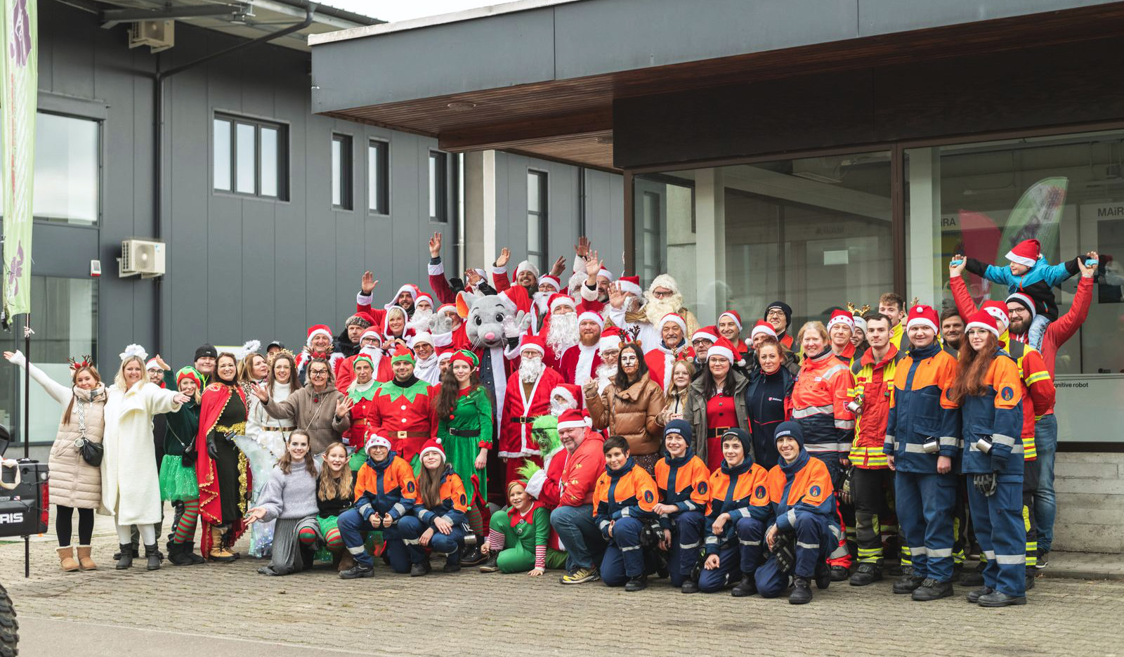 Große Gruppe von Menschen, viele verkleidet als Weihnachtsfiguren wie Santa Claus, Elfen und Rentieren, einige tragen orangeblaue Einsatzuniformen, versammelt vor einem modernen Gebäude und posieren lachend für ein Gruppenfoto.