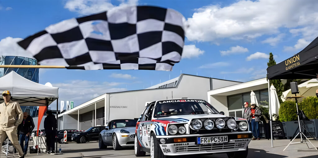 Rennwagen Lancia Martini mit hellblau-roten Rennstreifen fährt unter einer schwarz-weiß karierten Zielflagge in einem Motorsport-Event mit Zuschauern und Zelten im Hintergrund.