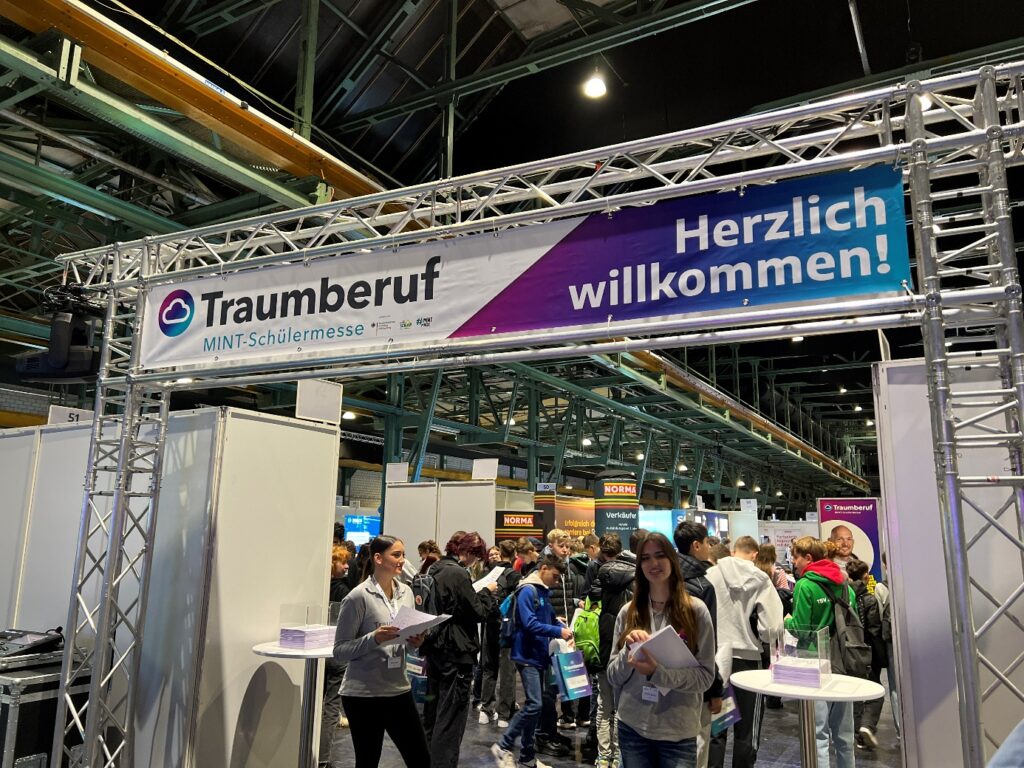 Messestand mit einem Banner "Traumberuf MINT-Schülermesse Herzlich willkommen!" und vielen jugendlichen Besuchern in einer großen Halle mit sichtbarer Stahlkonstruktion im Dachbereich