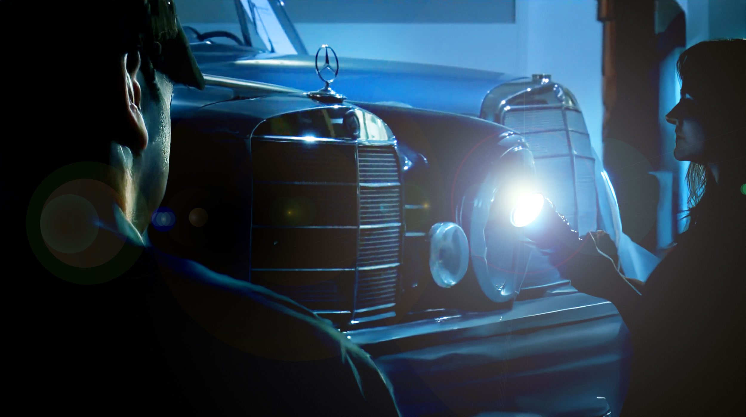 In a dark garage, two people use a flashlight to illuminate an old blue Mercedes-Benz with large headlights and the typical radiator grille.