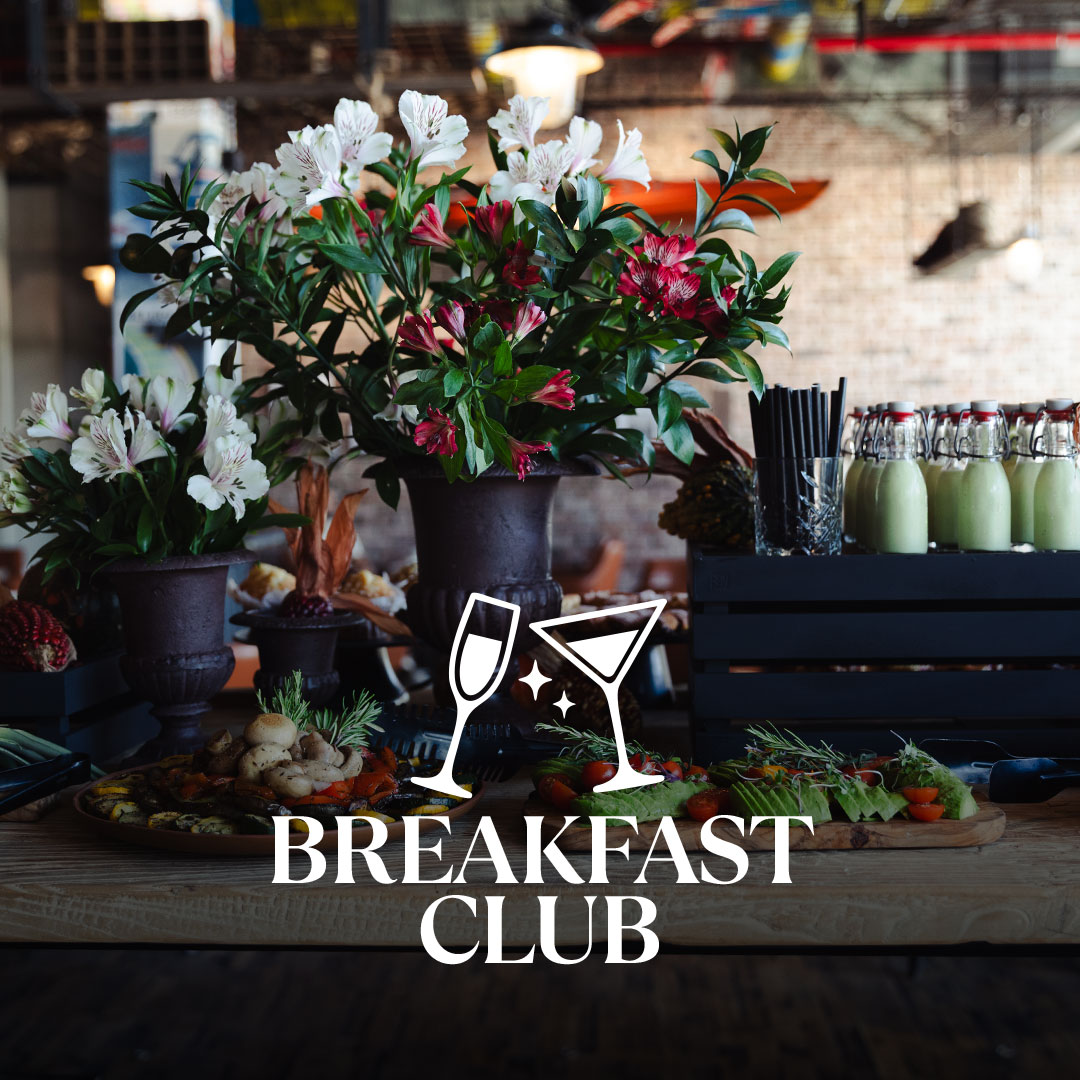 Rustikaler Holztisch in einem Café mit großen Vasen voller weißer und rosa Blumen, Platten mit Frühstücksleckereien und mehreren mit hellgrüner Flüssigkeit gefüllten Bügelflaschen; darüber ist ein weißes Logo mit zwei anstoßenden Gläsern und dem Schriftzug BREAKFAST CLUB eingeblendet.