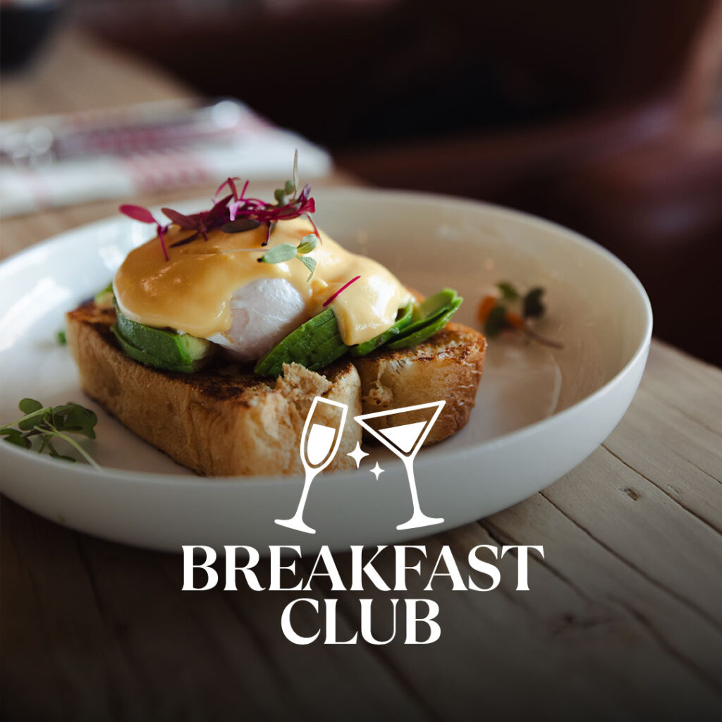 Eggs Benedict auf geröstetem Toast mit Avocado, pochiertem Ei und Hollandaise, garniert mit Mikrogrün in einer weißen Schale auf einem Holztisch, unten überlagert vom Schriftzug ‚BREAKFAST CLUB‘ und zwei Cocktailgläsern.