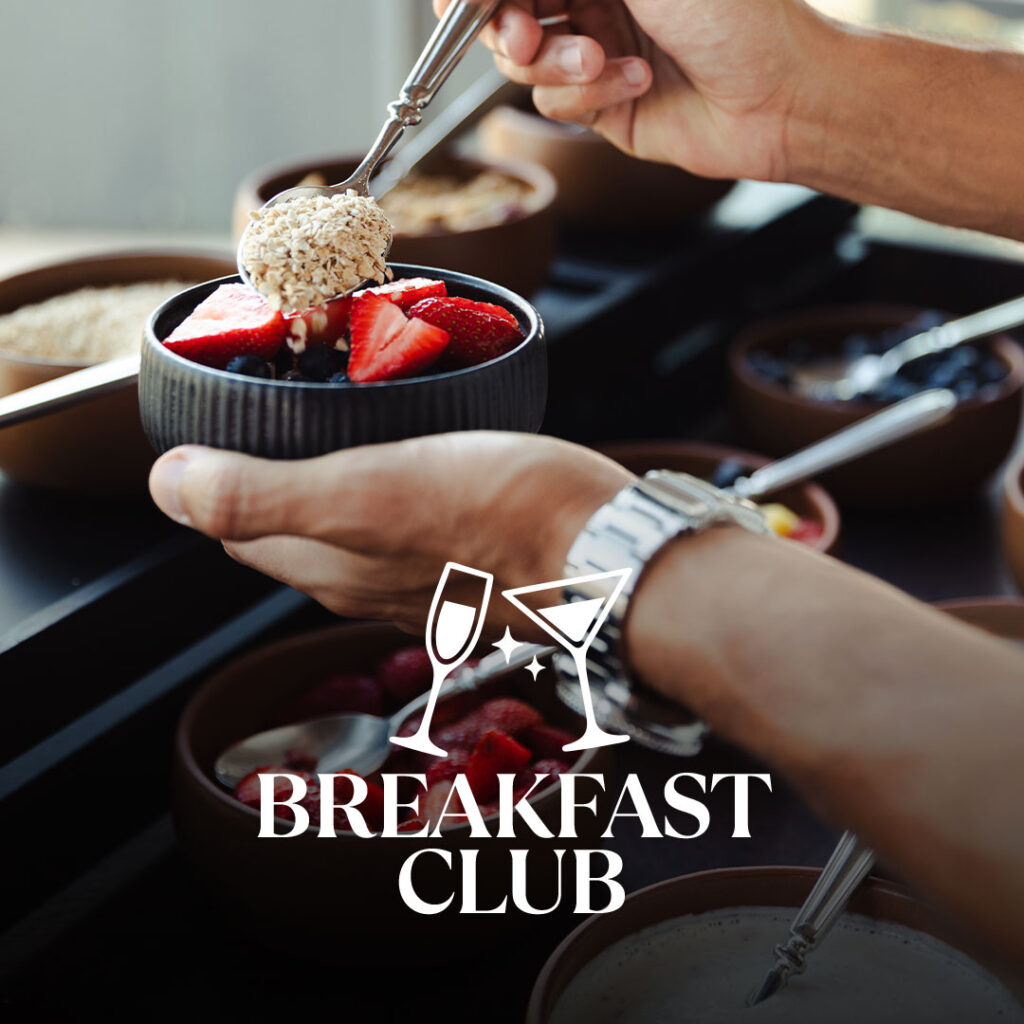 Unas manos sostienen un cuenco oscuro con rodajas de fresas y arándanos mientras se espolvorean copos de avena por encima con una cuchara; al fondo, varios cuencos pequeños y un logotipo blanco del "BREAKFAST CLUB" con vasos estilizados se superponen a la imagen.