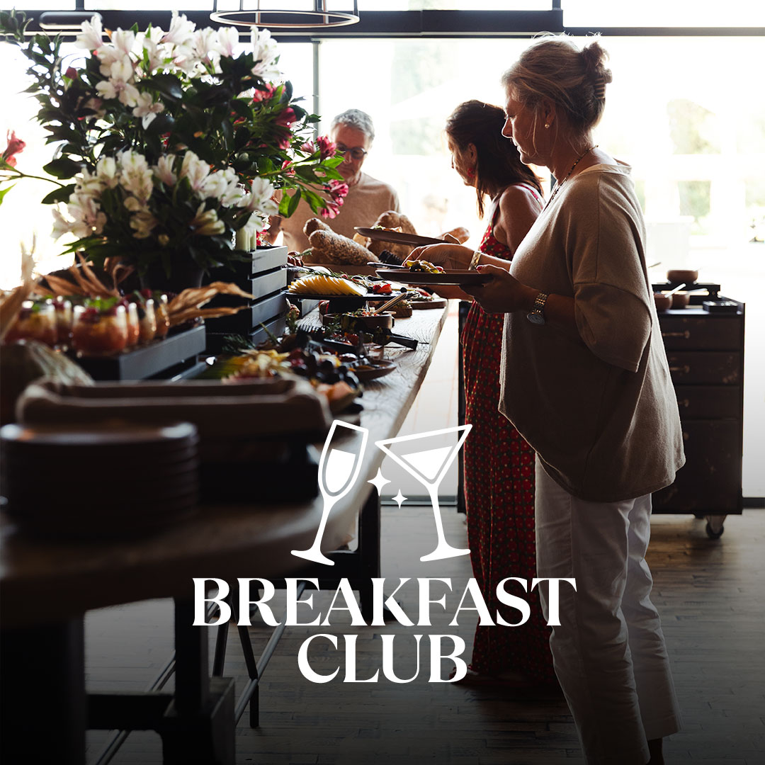 Drei Personen bedienen sich an einem reich bestückten Buffet mit großen Blumenarrangements in einem hellen Innenraum; im Vordergrund Teller und Speisen, unten überlagert das weiße Logo „BREAKFAST CLUB“ mit Cocktailgläsern.