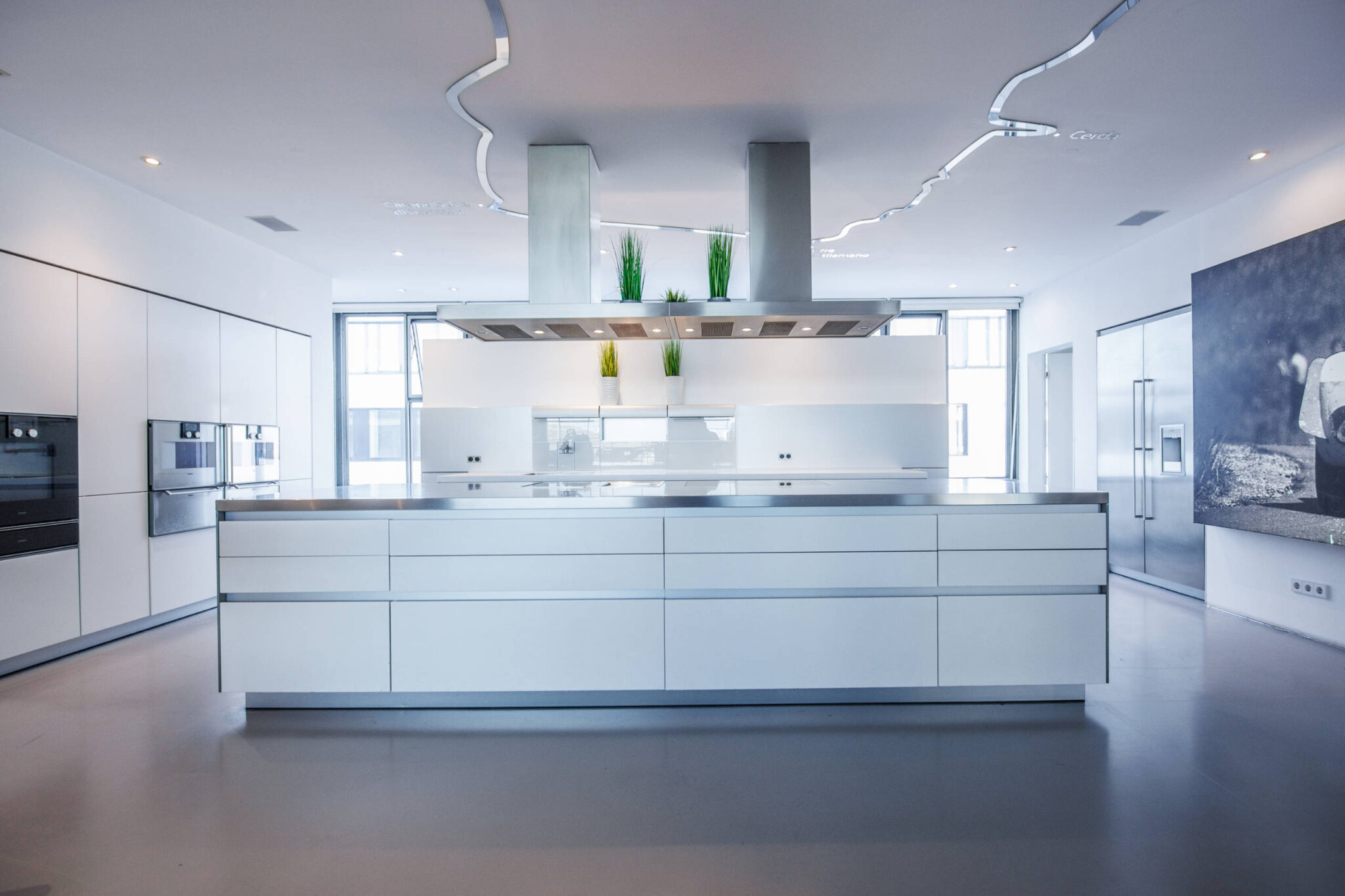 Moderne, minimalistische weiße Küche mit großer zentraler Insel und Edelstahlarbeitsplatte, zwei deckenmontierten Dunstabzugshauben, eingelassenen Geräten, drei kleinen Grünpflanzen auf einem Regal, großen Fenstern und geschwungener Deckenleuchte in einem hellen, offenen Raum.