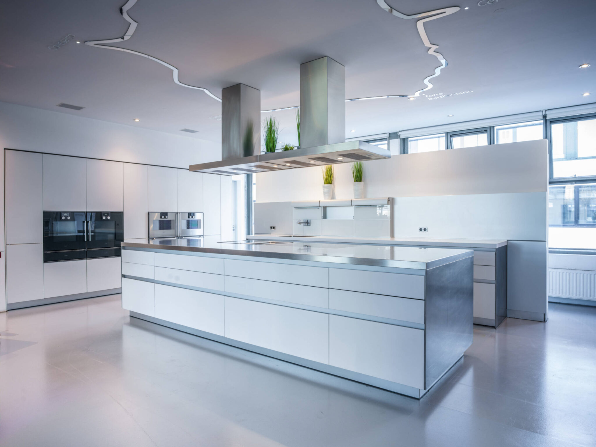 Helle, minimalistische Profi‑Küche mit großer zentraler Insel und Edelstahlarbeitsplatte, zwei markanten Edelstahl-Dunstabzügen darüber, weiße Hochschränke mit eingebauten Öfen links, große Fenster rechts, geschwungene Deckenbeleuchtung und drei kleine grüne Topfpflanzen als Farbakzente.