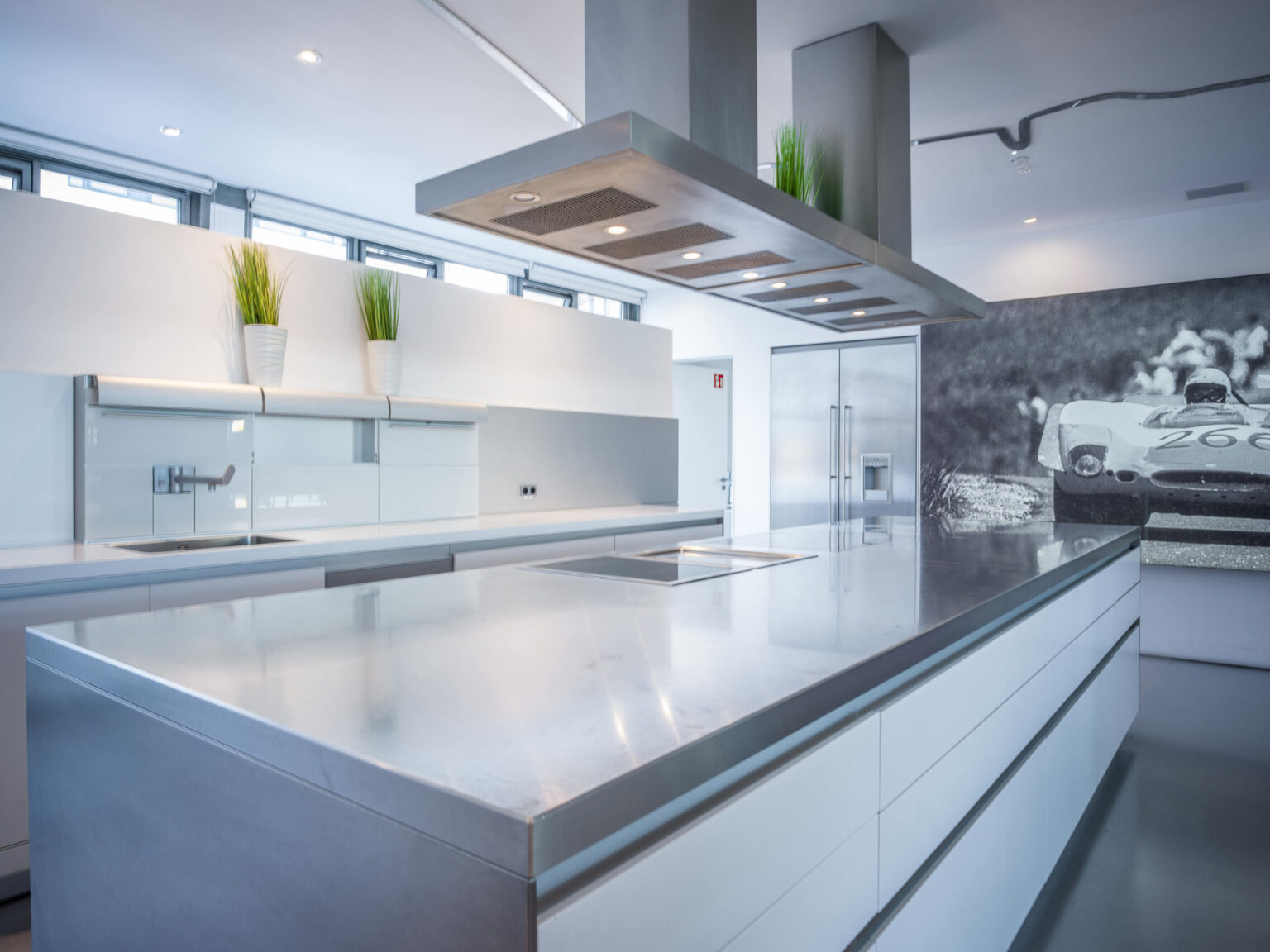 Moderne weiße Küche mit großer Edelstahl-Insel, überhängender Dunstabzugshaube und Retro-Rennwagen-Wandbild.