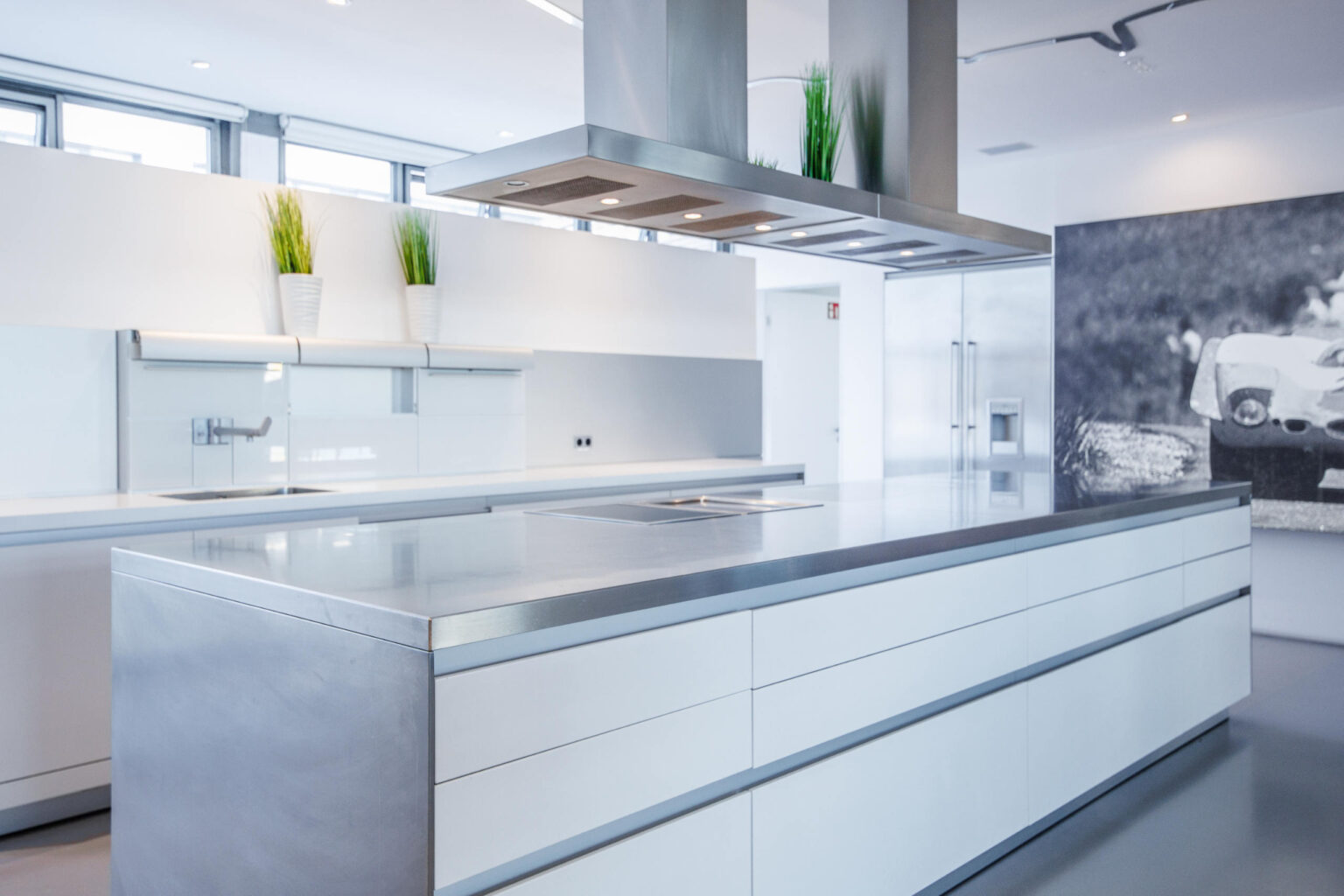 Minimalistische weiße Küche mit großer Insel, Edelstahlarbeitsplatte, Dunstabzug und zwei Topfpflanzen.