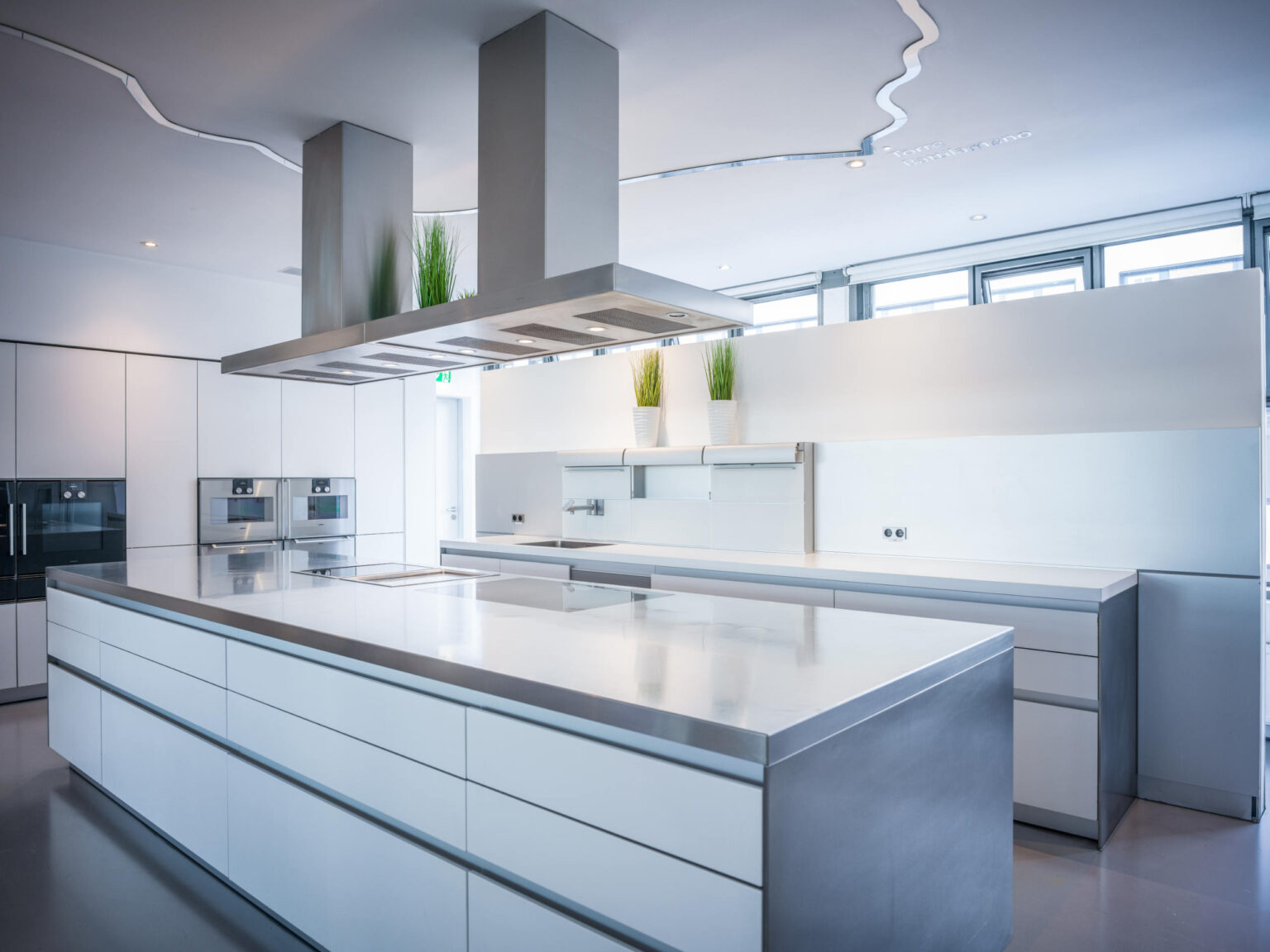 Moderne minimalistische weiße Küche mit großer Mittelinsel, Edelstahl-Dunstabzug und eingebauten Geräten.
