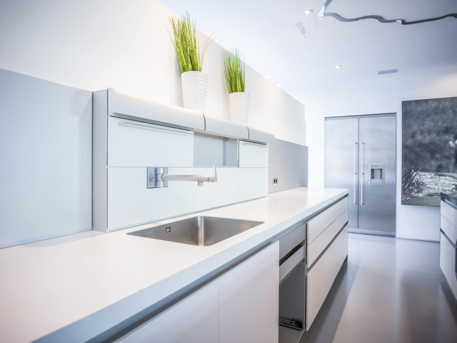 Helle minimalistische Küche mit weißer Arbeitsplatte, integriertem Edelstahlspülbecken und -armatur, großem Edelstahl-Kühlschrank und zwei hohen Pflanzen.