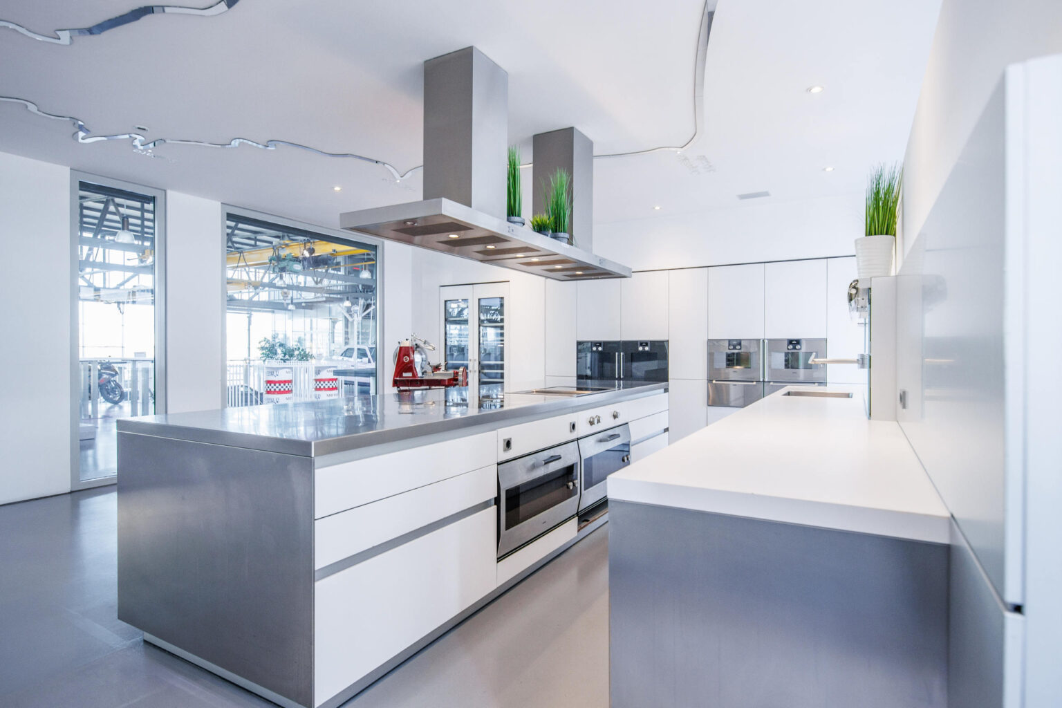 Moderne helle Küche mit großer Edelstahl-Kochinsel, eingebauten Öfen, großer Dunstabzugshaube und dezenten Pflanzen.