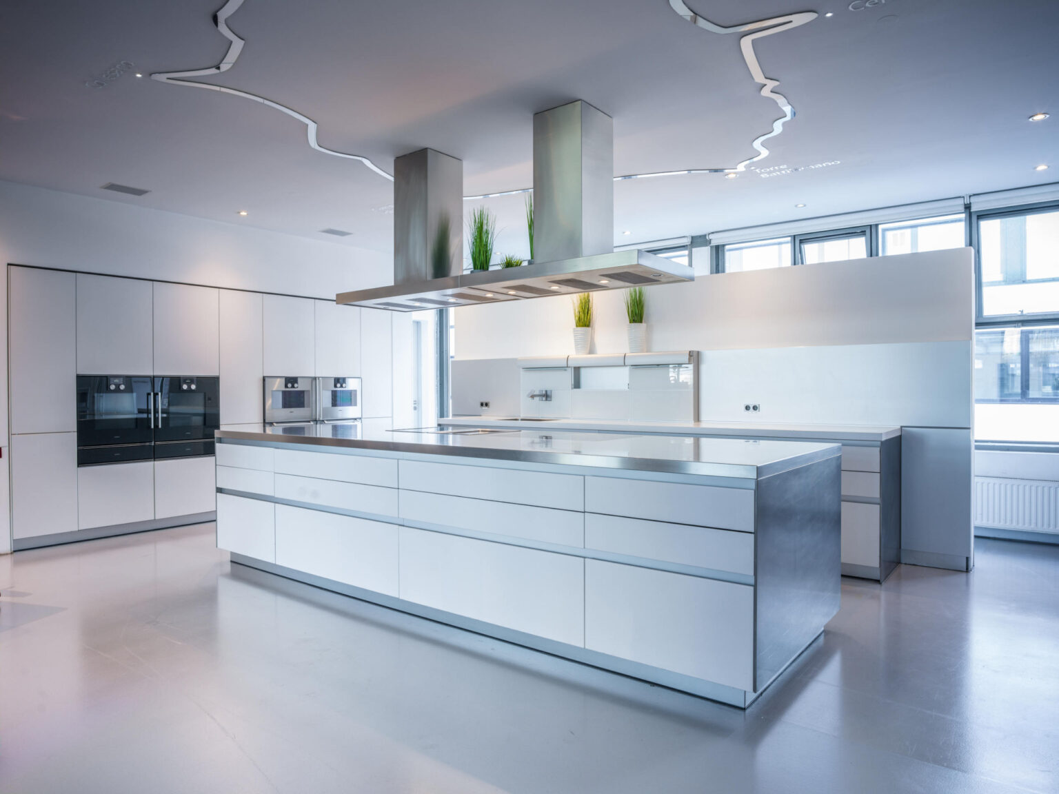 Helle, minimalistische Küche mit großer weißer Insel, Edelstahlarbeitsfläche, Dunstabzug und eingebauten Geräten