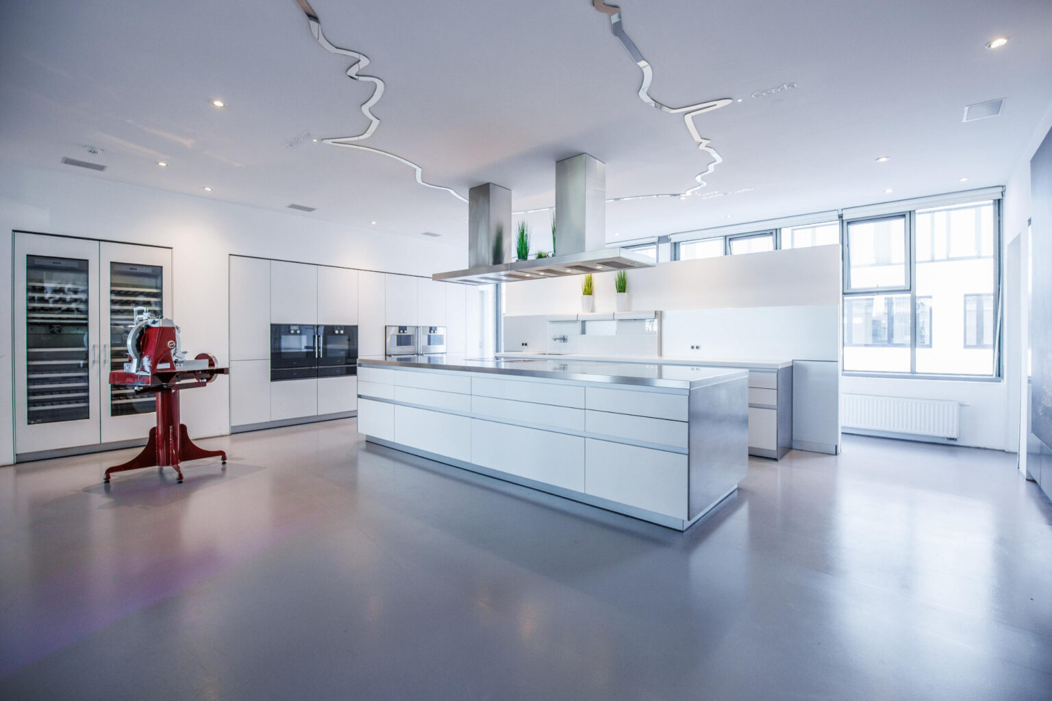 Große weiße Profi-Küche mit zentraler Insel, Edelstahl-Dunstabzugshauben und roter Aufschnittmaschine links.