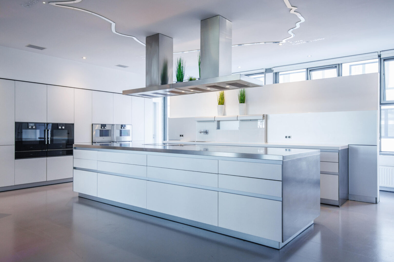 Moderne minimalistische weiße Küche mit großer Mittelinsel, Edelstahl-Dunstabzugshaube und eingebauten Geräten.