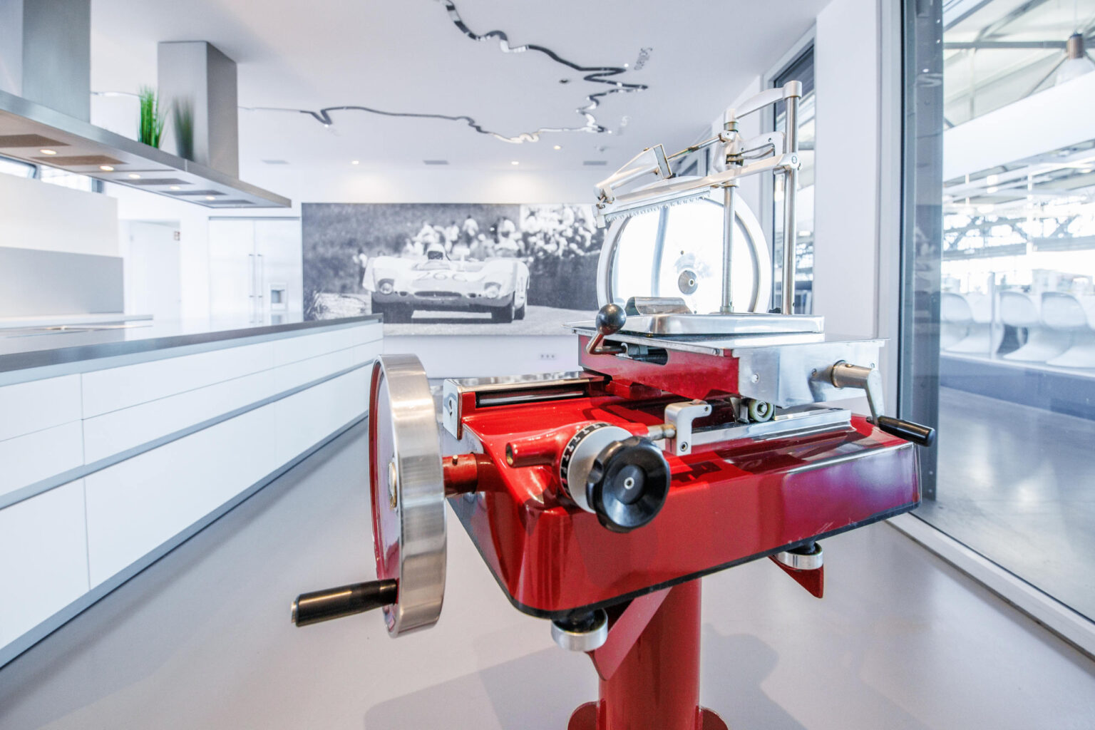 Rote Retro-Aufschnittmaschine mit großem Metallrad in hellem modernen Raum vor Schwarzweißfoto eines Rennwagens.