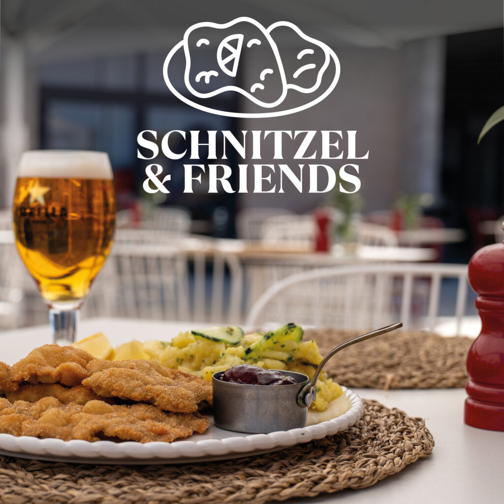 A laid table with a plate of several breaded schnitzels, a small metal bowl with cranberry sauce, potato salad with cucumber slices and lemon wedges, a glass of beer on the left and a red pepper mill on the right; in the blurred background an outdoor café area and the white logo 'SCHNITZEL & FRIENDS' at the top.