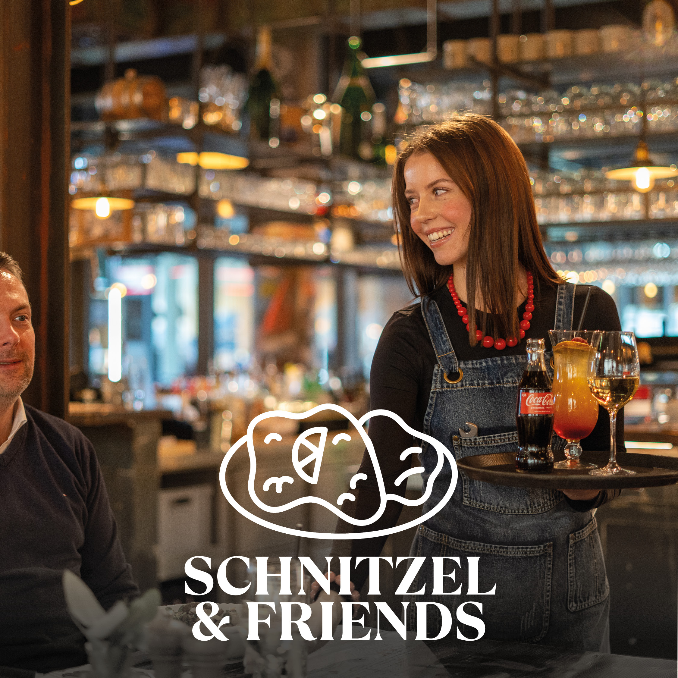 Young waitress in denim dungarees and red pearl necklace carries a tray with a Coca-Cola bottle, a cocktail and a wine glass in a warmly lit restaurant, a guest sits on the left and the logo 'Schnitzel & Friends' is superimposed on the front with a stylized schnitzel graphic.