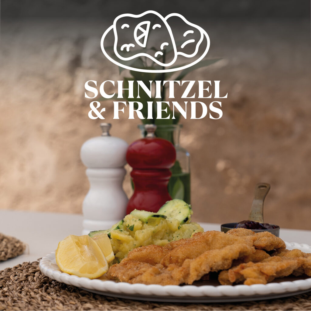 Foreground: plate with crispy breaded schnitzel, lemon wedges and potato salad; in the background salt and pepper mills, glass vase and above it the white logo "SCHNITZEL & FRIENDS" with stylized schnitzel illustration.