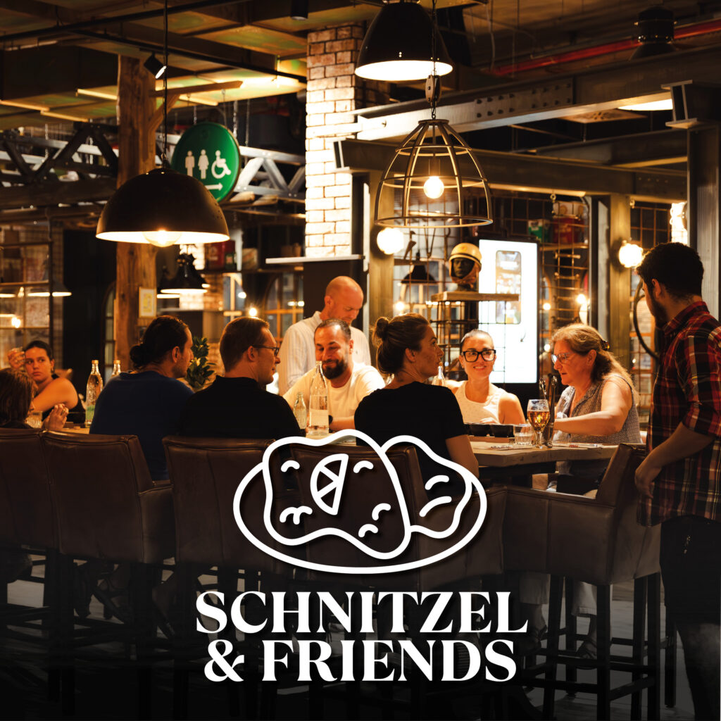 Mehrere Gäste sitzen unter hängenden Lampen in einer warm beleuchteten, industriell gestalteten Bar/Restaurant und unterhalten sich; unten mittig ein weißes Logo mit stilisierten Schnitzeln und dem Schriftzug „Schnitzel & Friends“.