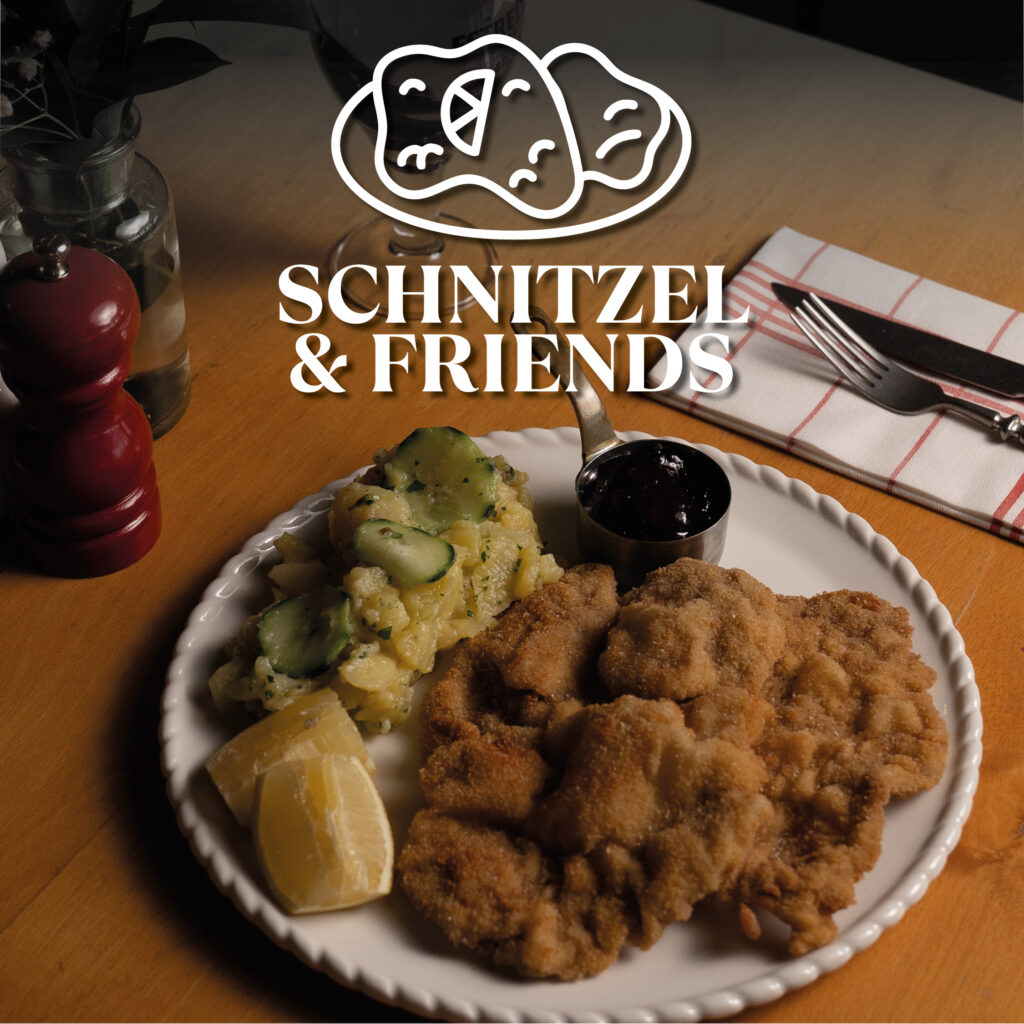 Auf einem Holztisch ein weißer Teller mit knusprigen panierten Schnitzeln, Kartoffelsalat mit Gurkenscheiben, Zitronenspalten und einem kleinen Becher dunkler Beilage, rechts Besteck auf kariertem Stoff, links eine rote Pfeffermühle und oben über dem Motiv das weiße Logo „SCHNITZEL & FRIENDS“.