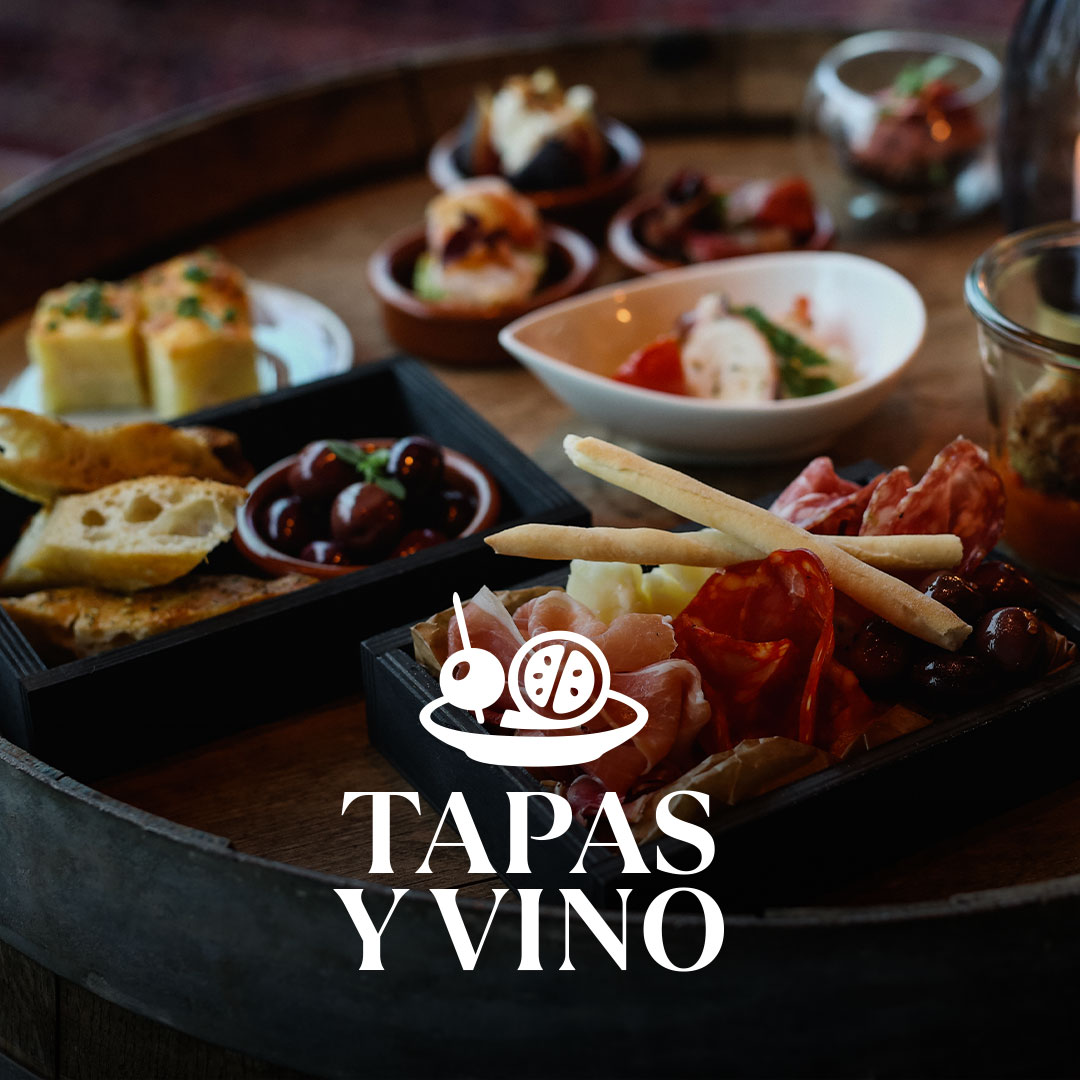 Verschiedene Tapas in kleinen Schälchen und Holztabletts (Brot, Oliven, Aufschnitt, Brotstangen und Käse) auf einem runden Fass als Tisch, mit weißem Schriftzug „TAPAS Y VINO“ im Vordergrund.