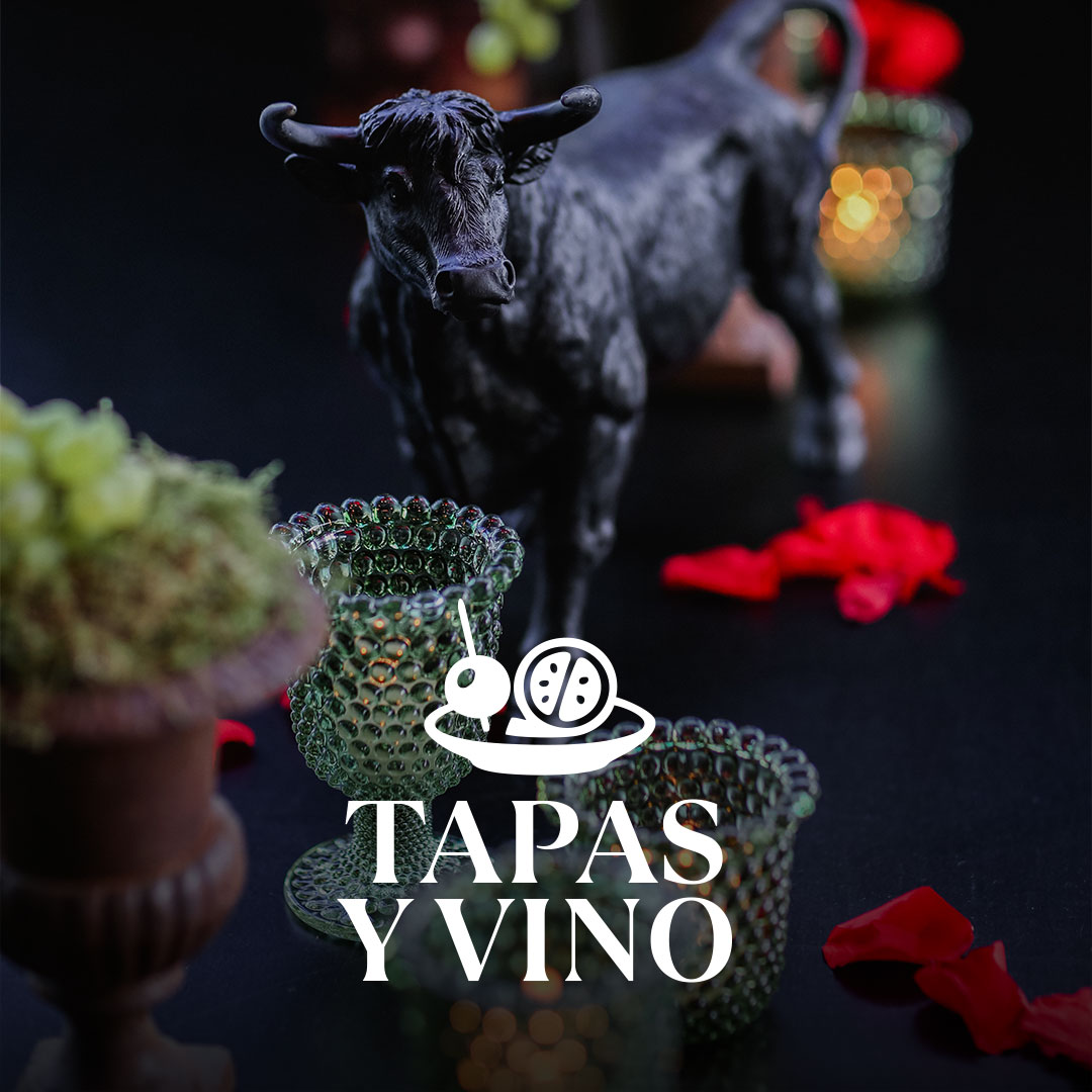 Schwarze Stierfigur auf dunklem Tisch, davor grün strukturierte Glasgefäße, rote Rosenblätter und ein kleines Pflanzgefäß; mittig ein weißes Logo mit Oliven-auf-Teller und dem Schriftzug TAPAS Y VINO.