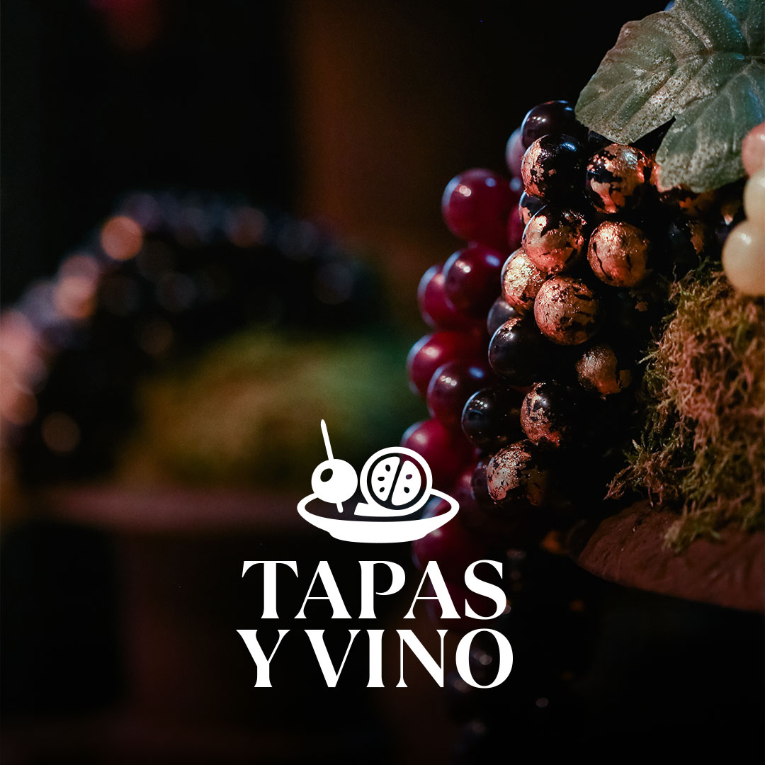 Nahaufnahme eines dunklen, teilweise goldbemalten Traubenbündels mit Blatt vor unscharfem Hintergrund und dem weißen Schriftzug TAPAS Y VINO samt kleiner Teller- und Tapas-Ikone im Vordergrund.