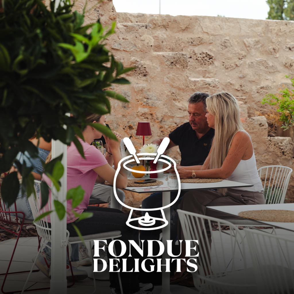 Drei Personen sitzen an einem Außentisch vor einer Steinmauer, darüber Logo FONDUE DELIGHTS.