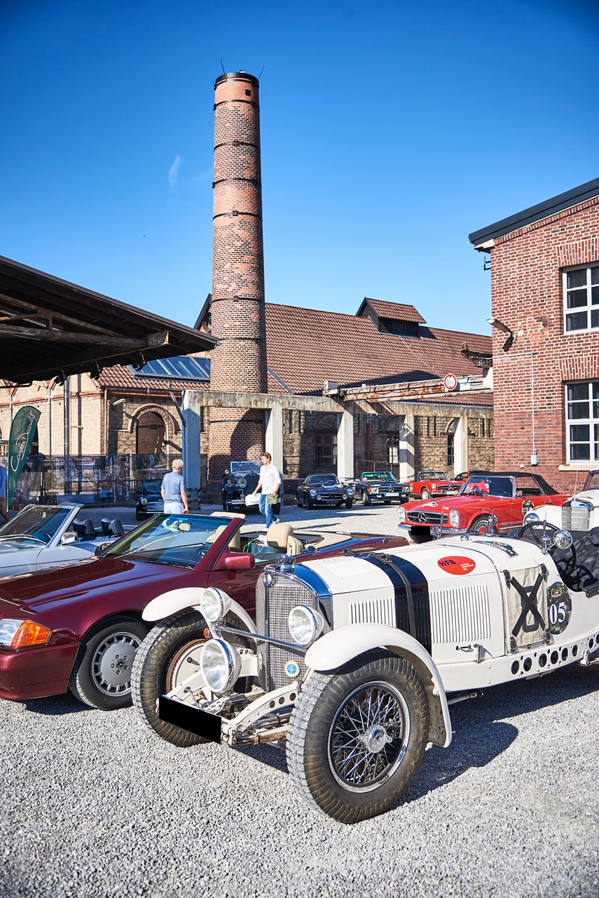 Historischer weißer Rennwagen im Vordergrund, weitere Oldtimer auf Hof vor roter Backsteinfabrik mit hohem Schornstein.