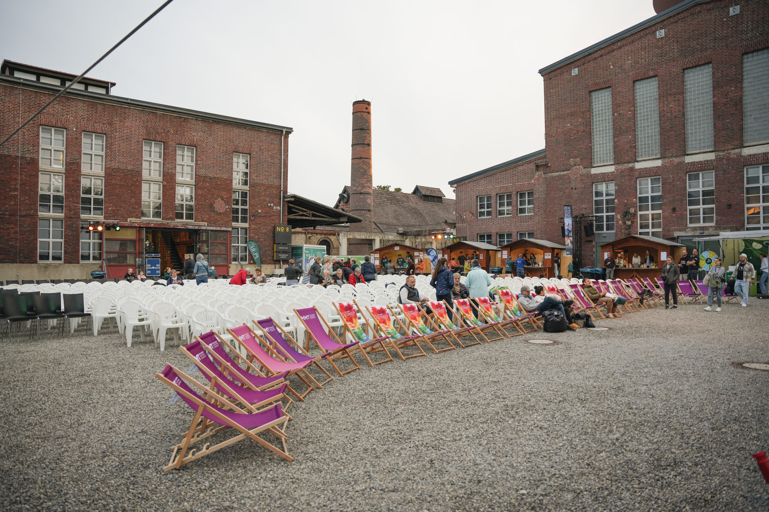 Reihen bunter Liegestühle und weißer Plastikstühle in einem Industrieinnenhof vor Backsteingebäuden mit Schornstein und Besuchern.