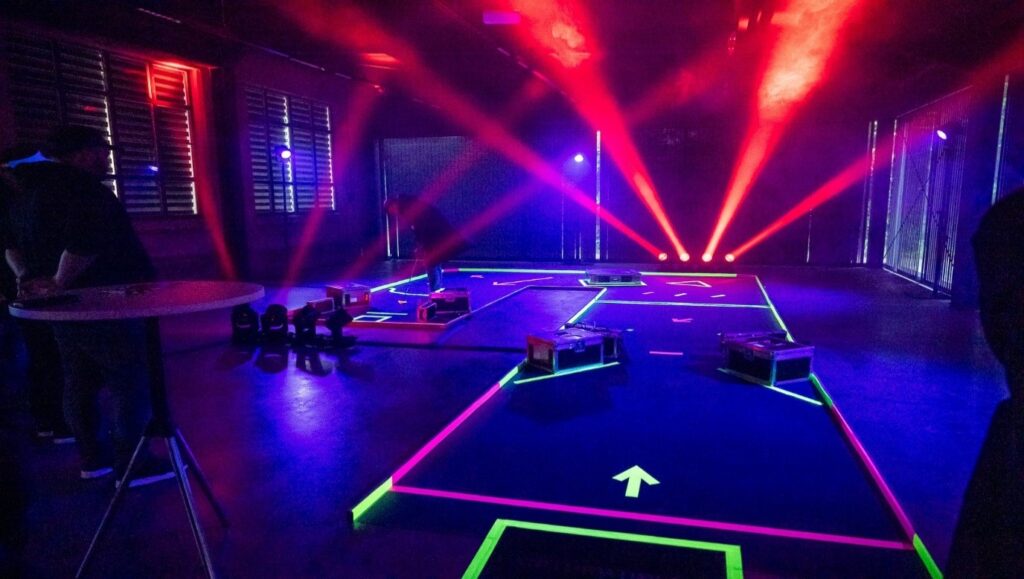 Dunkler Innenraum mit leuchtenden Neon-Pisten auf dem Boden und roten Bühnenlichtern, die in Rauch strahlen.